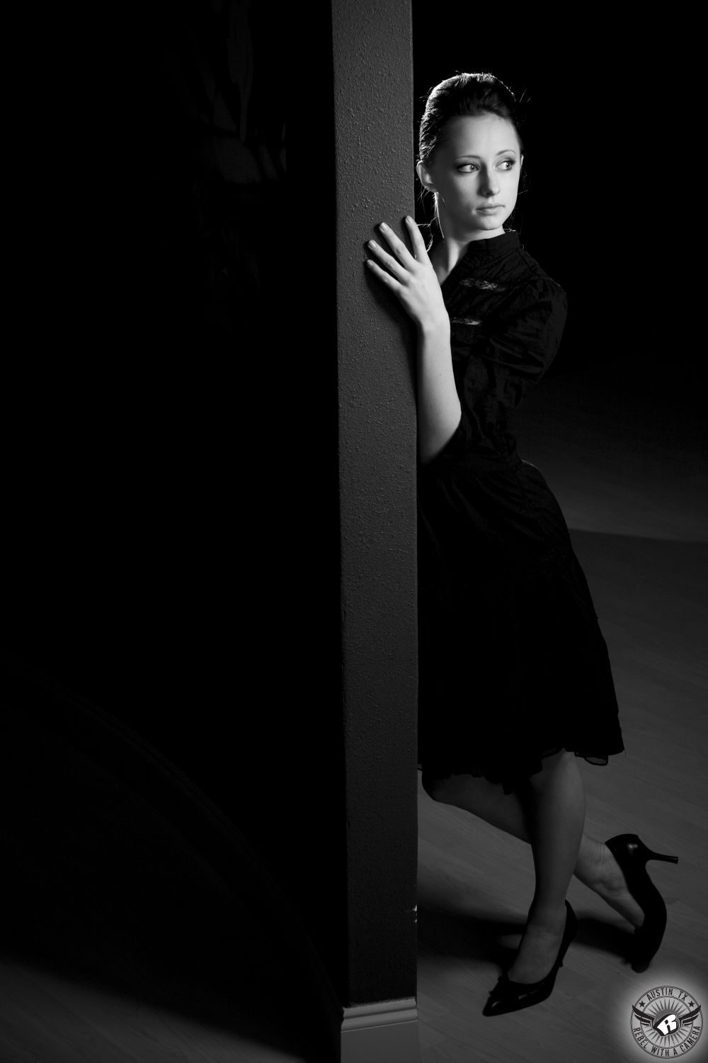 sultry brunette fashion female model  in a black dress with black four inch heals softly lit leaning against a wall with her toe pointed into the wooden floor